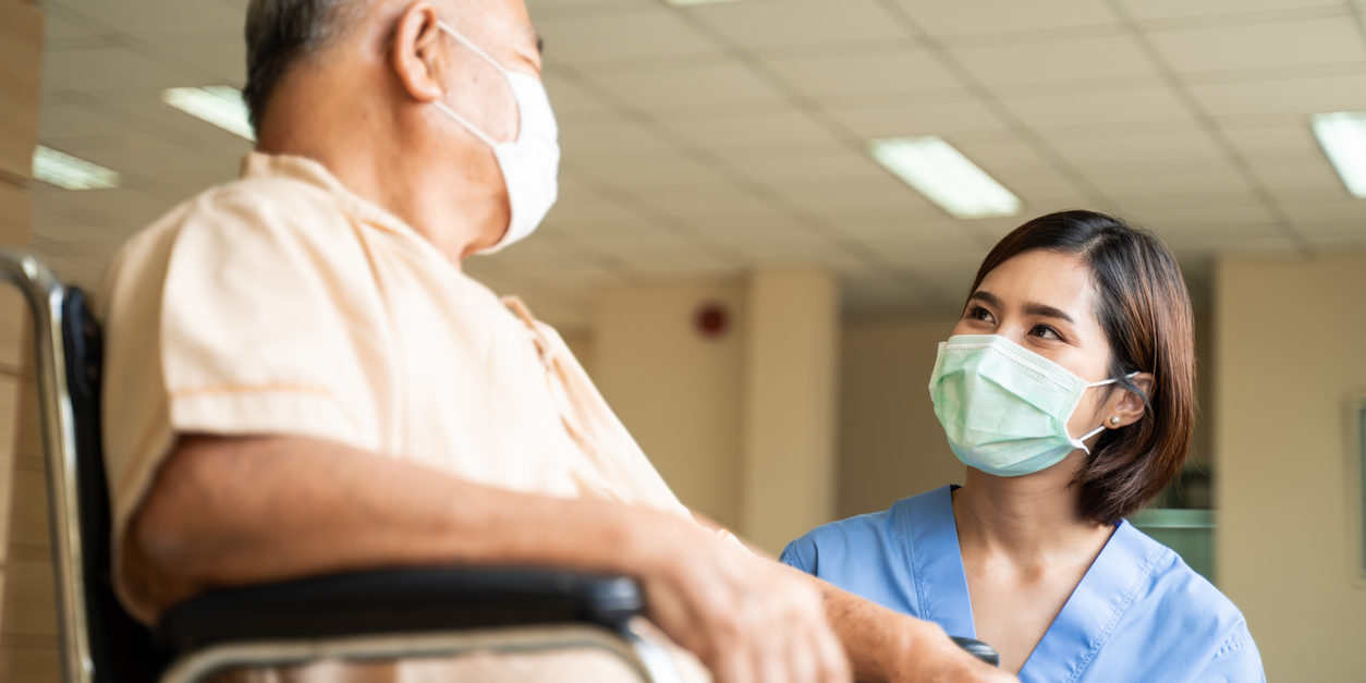 Asian nurse taking care of mature male patient sitting on wheelchair in hospital. Young woman and old man wearing surgical face mask for protection of The COVID pandemic. Girl talking to elderly man.